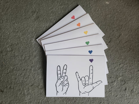 Shown is a pack of 6 white cards that have been block printed with two hands, one showing peace and the other I love you. Above the love hand is a tiny heart, each card has a different color of the rainbow heart. 